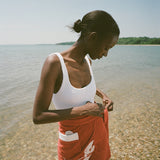 woman at beach wearing white one piece bathing suit and red skirt, looking down and tying wrap on her skirt