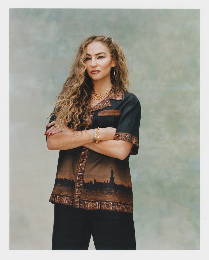 Female model with long blonde hair and crossed arms wearing 'The Sopranos' Turnpike Batik and black pants, shot on a light green background.