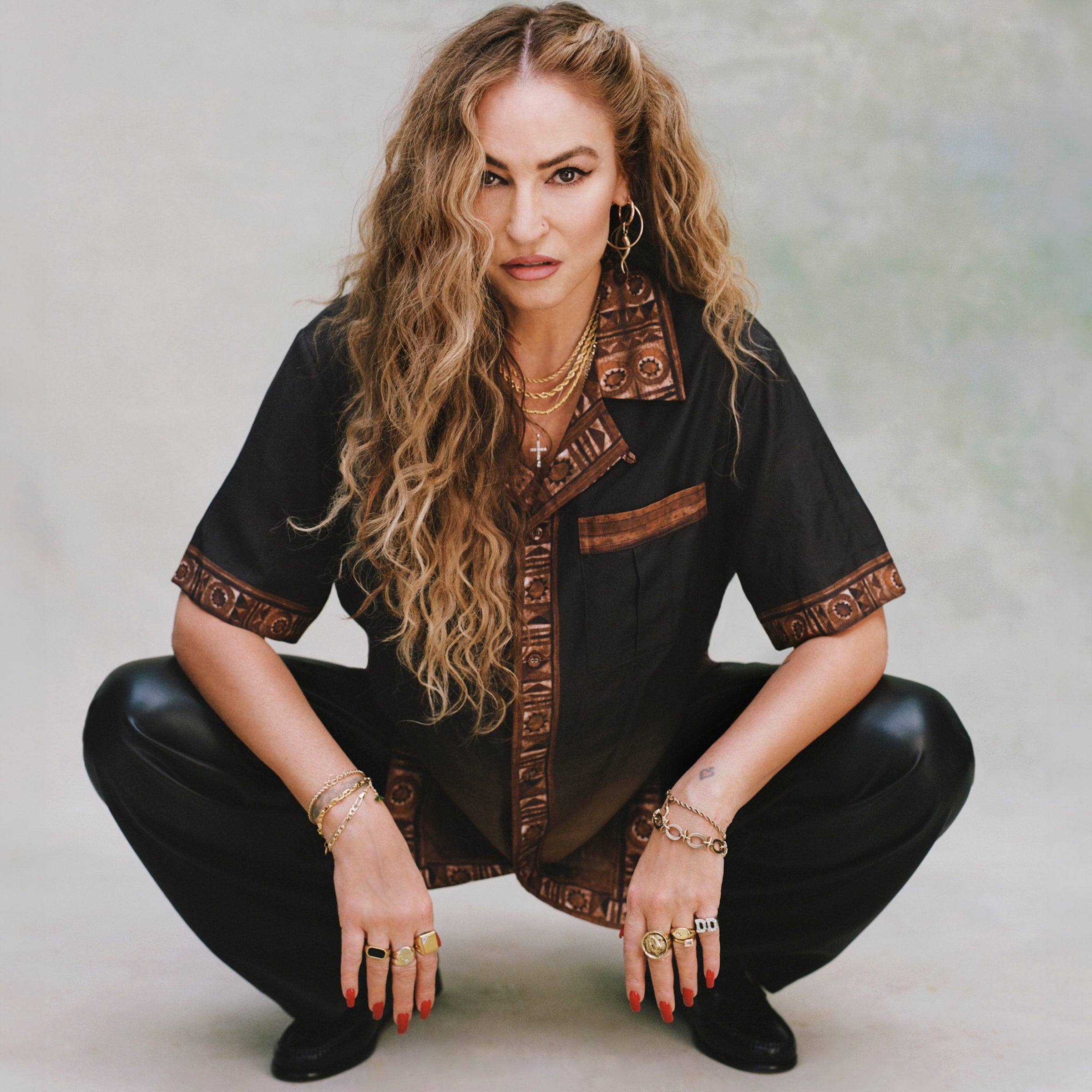 Female model with long blond hair in a crouching position wearing 'The Sopranos' Turnpike Batik with digitally printed NYC skyline, contrasting batik collar, cuffs, placket, and bottom sweep, while showcasing its double chest pockets with concealed interior buttons.