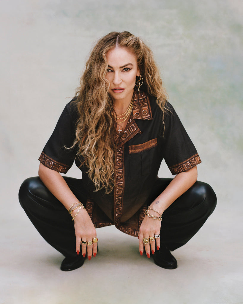 Female model with long blond hair in a crouching position wearing 'The Sopranos' Turnpike Batik with digitally printed NYC skyline, contrasting batik collar, cuffs, placket, and bottom sweep, while showcasing its double chest pockets with concealed interior buttons.