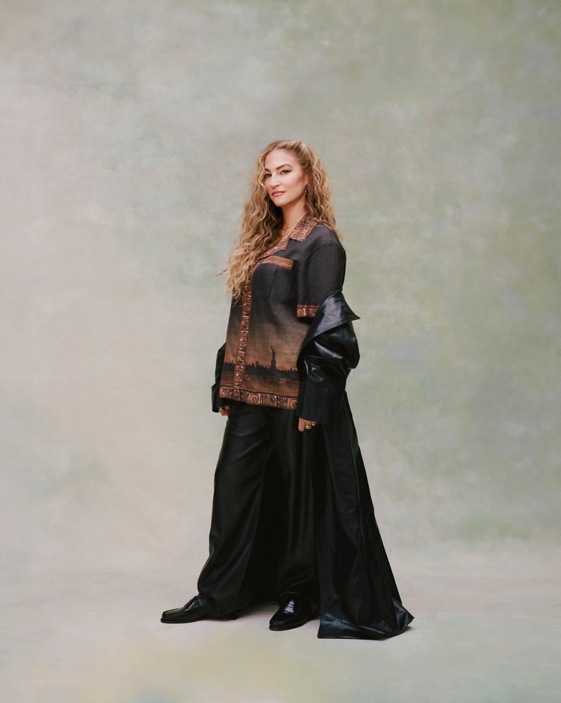 Female model with long blond hair standing to the left with her faced turned toward the camera wearing 'The Sopranos' Turnpike Batik with black flowy pants, black shoes, and black leather overcoat being worn half on or off by the model, shot on a light green background.