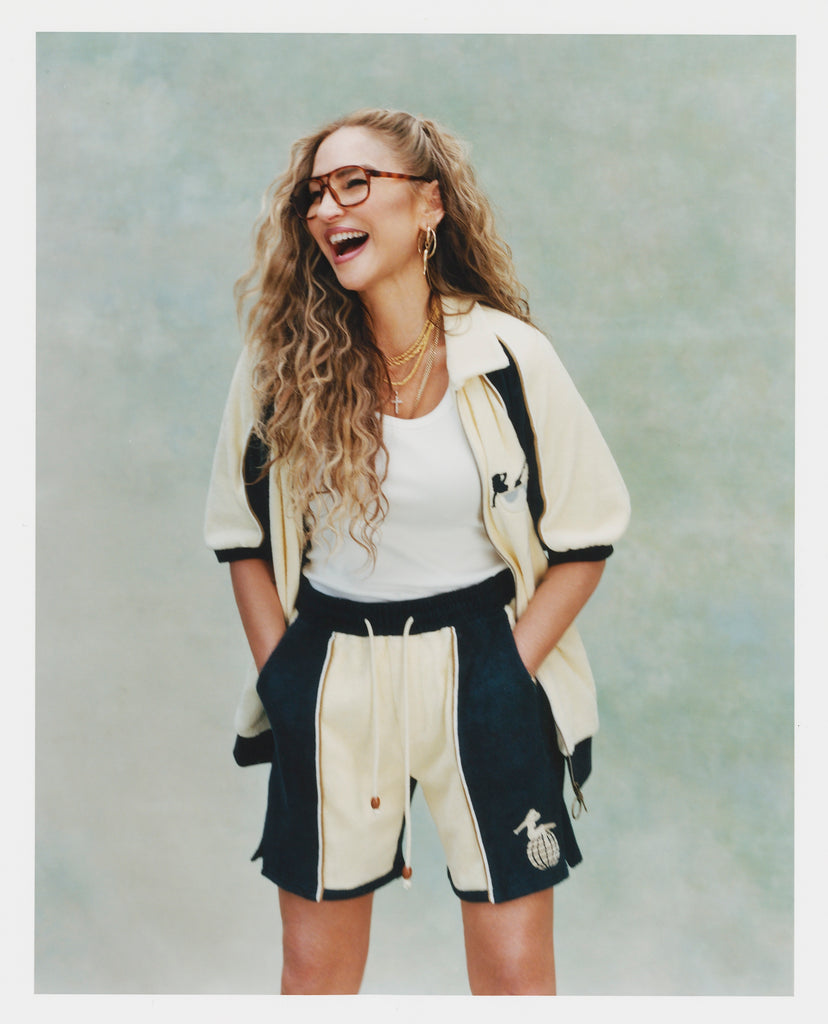 Female model with long blonde hair wearing 'The Sopranos' Bada Bing Cabana unbuttoned with a white shirt, matching Bada Bing shorts, and glasses, laughing, shot on a textured light blue background.