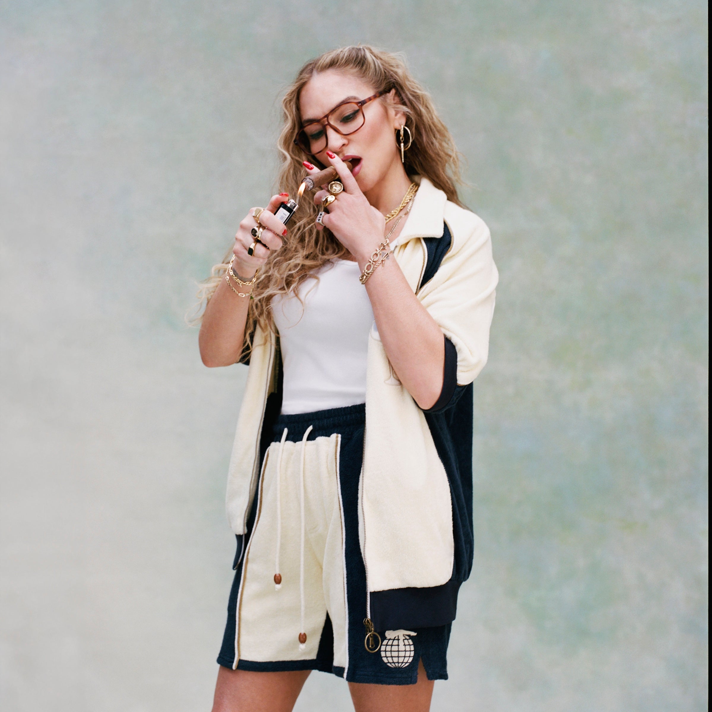Female model with long blonde hair lighting a cigar while wearing 'The Sopranos' Bada Bing Cabana Shorts with an unbuttoned matching Bada Bing cabana and white shirt, shot a textured white background.
