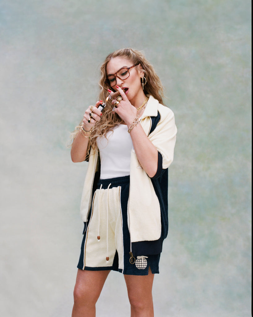 Female model with long blonde hair lighting a cigar while wearing 'The Sopranos' Bada Bing Cabana Shorts with an unbuttoned matching Bada Bing cabana and white shirt, shot a textured white background.