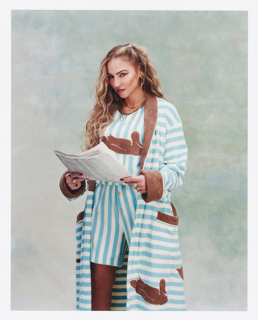 Female model with long blonde hair holding a newspaper while wearing 'The Sopranos' Fowl Play Shorts in light blue and white striped coloring with matching Fowl Play shirt and robe, shot on a textured light blue background.