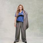 Female model with long curly blond hair wearing 'The Sopranos' Vesuvio Cabana ~ Crop Cut while holding a grey and white striped suit in one hand and wearing a matching grey and white-striped baggy pants and black loafers on a very light green background.