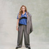 Female model with long curly blond hair wearing 'The Sopranos' Vesuvio Cabana ~ Crop Cut while holding a grey and white striped suit in one hand and wearing a matching grey and white-striped baggy pants and black loafers on a very light green background.