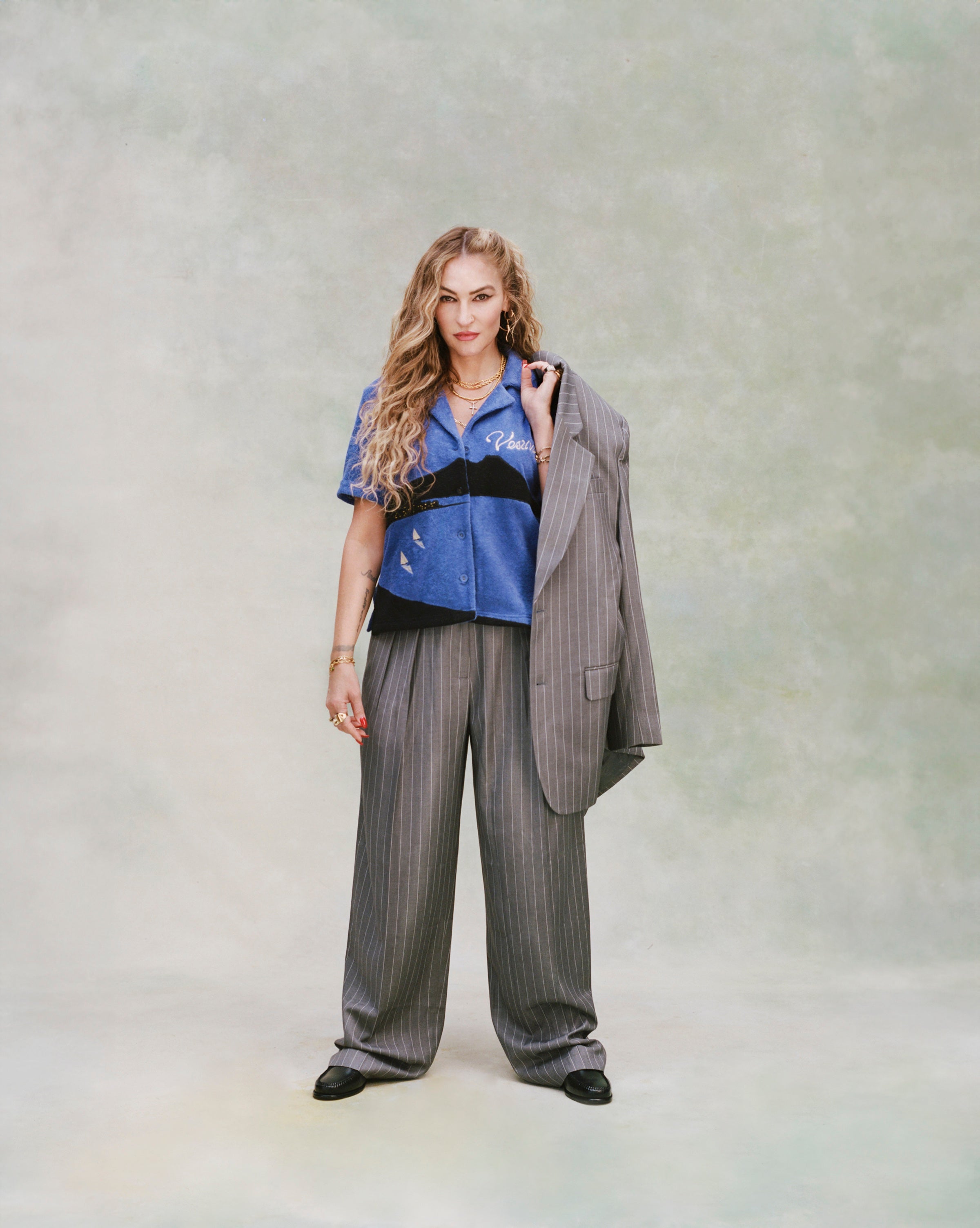 Female model with long curly blond hair wearing 'The Sopranos' Vesuvio Cabana ~ Crop Cut while holding a grey and white striped suit in one hand and wearing a matching grey and white-striped baggy pants and black loafers on a very light green background.