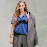 Female model with curly blonde hair and gold necklaces wearing 'The Sopranos' Vesuvio Cabana ~ Crop Cut while holding a grey and white striped blazer over her shoulder and wearing a matching grey and white-striped set of suit pants on very light green background. 