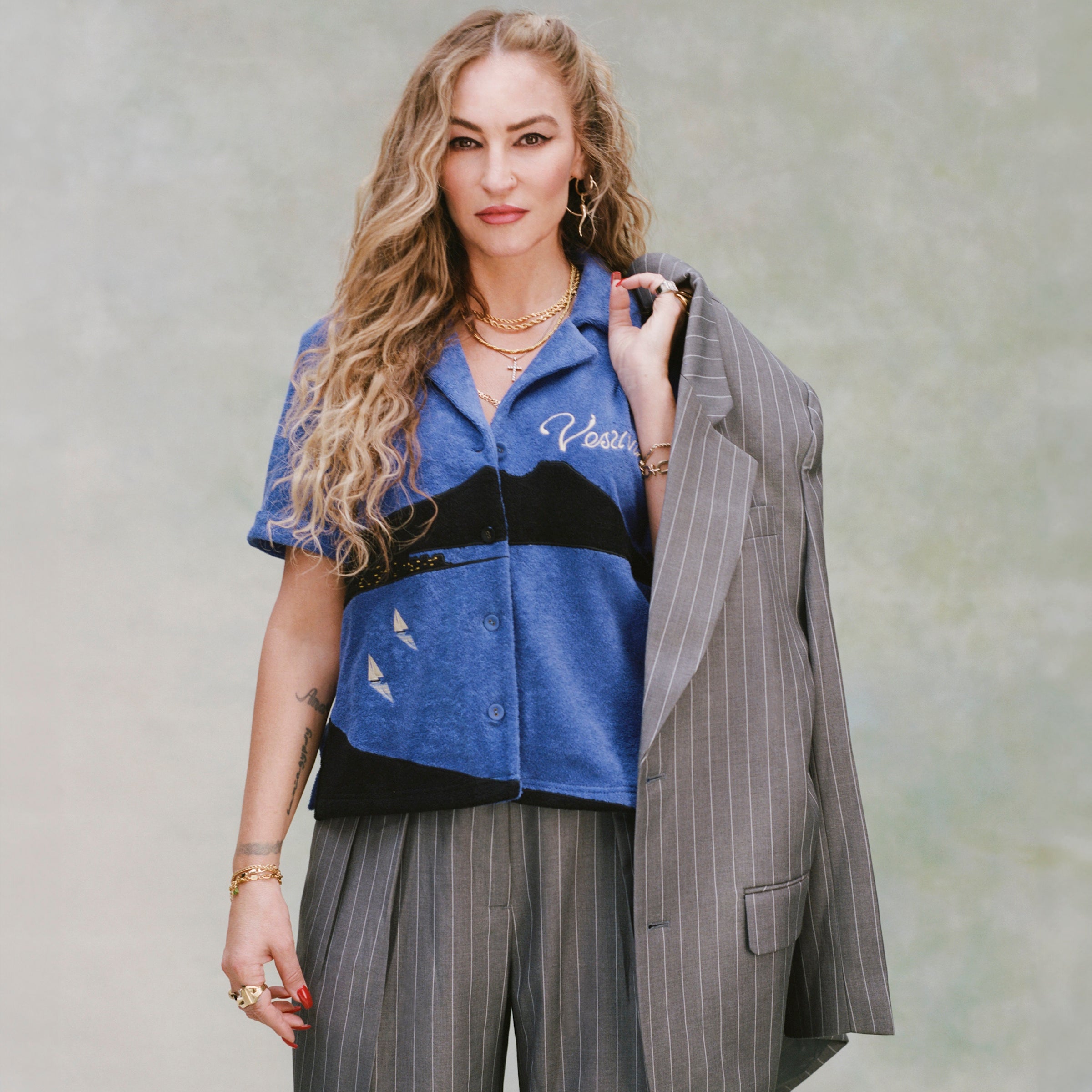 Female model with curly blonde hair and gold necklaces wearing 'The Sopranos' Vesuvio Cabana ~ Crop Cut while holding a grey and white striped blazer over her shoulder and wearing a matching grey and white-striped set of suit pants on very light green background. 
