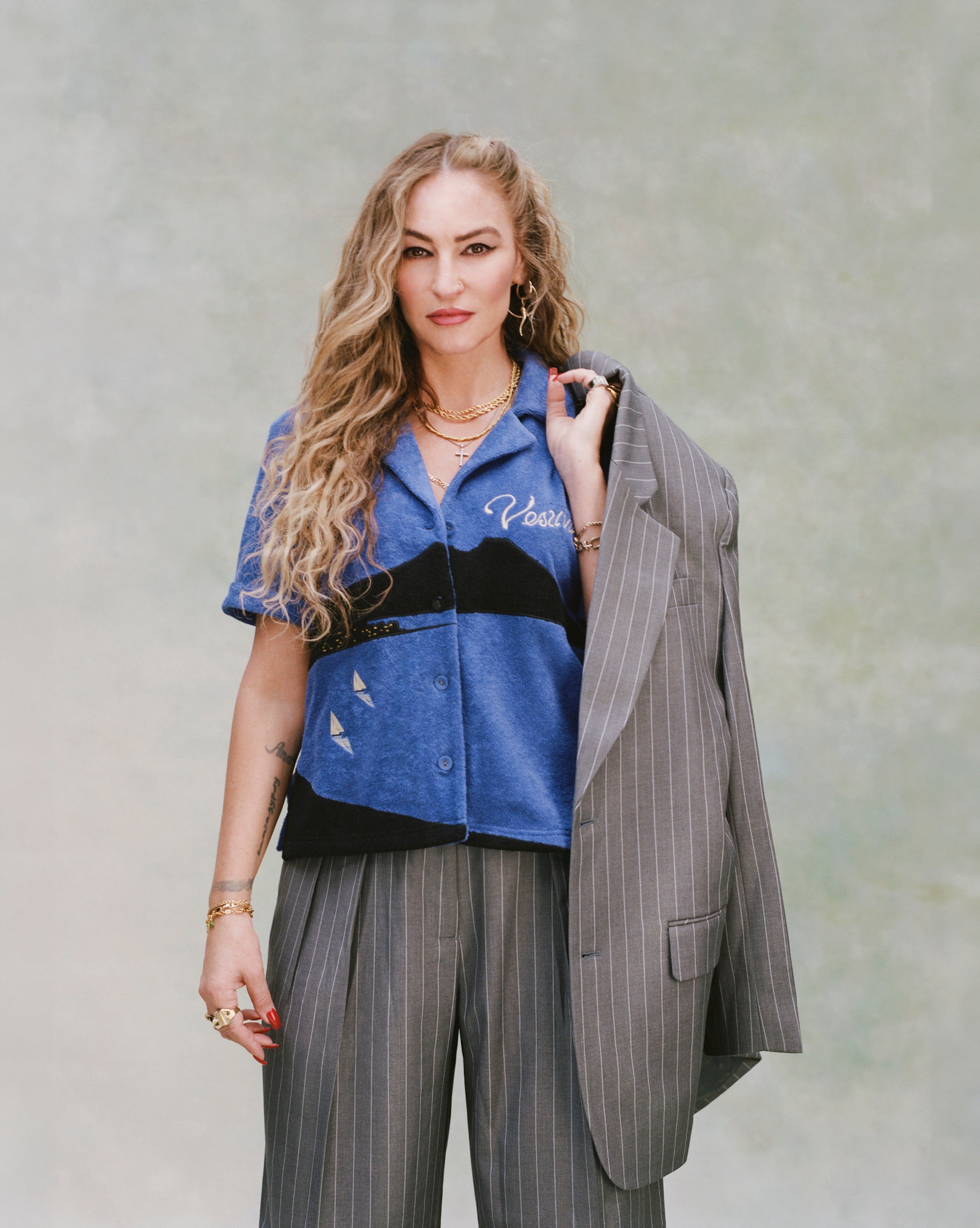 Female model with curly blonde hair and gold necklaces wearing 'The Sopranos' Vesuvio Cabana ~ Crop Cut while holding a grey and white striped blazer over her shoulder and wearing a matching grey and white-striped set of suit pants on very light green background. 