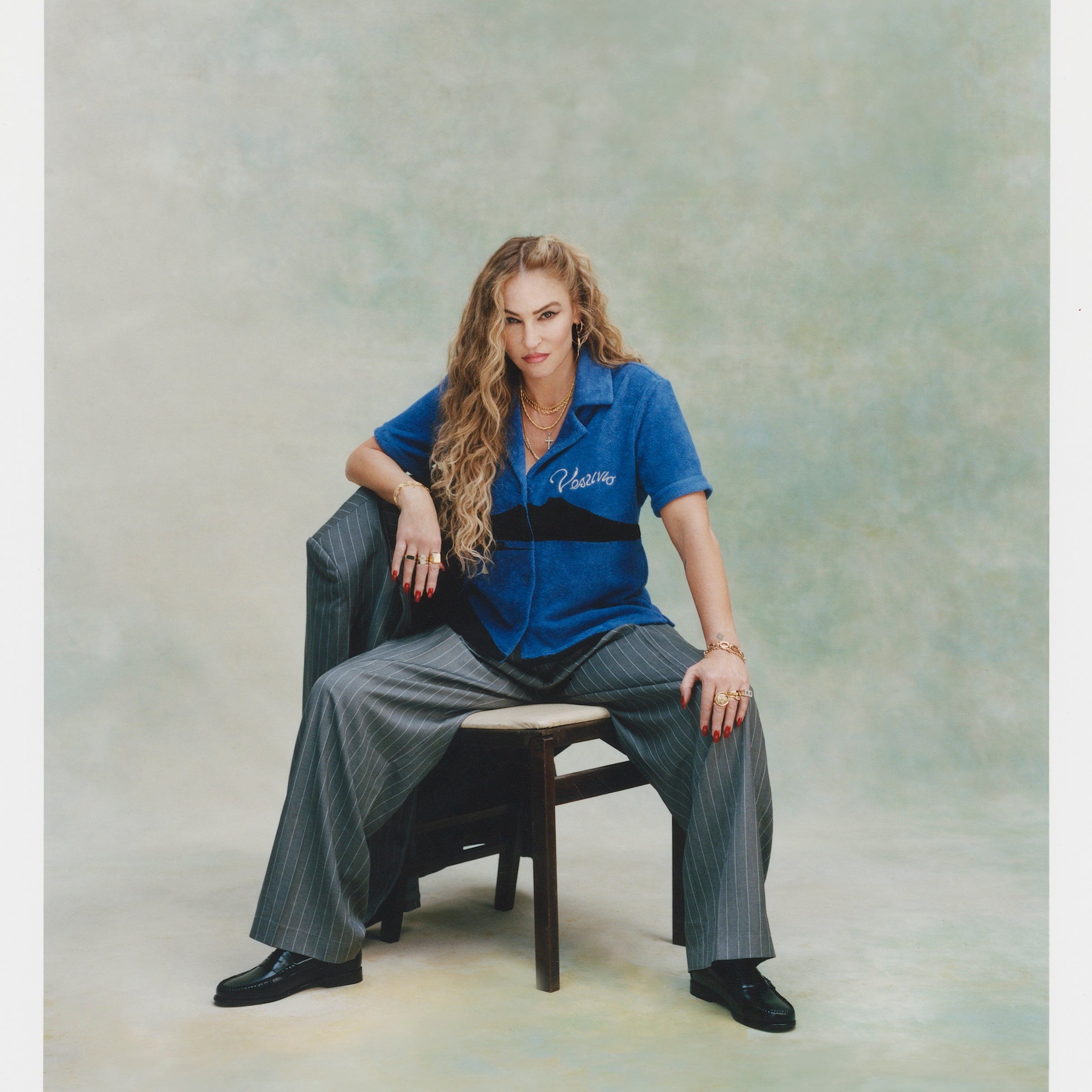 Female model with curly long blonde hair sitting facing the camera with one hand on her knee and the other resting against the backside of the chair in 'The Sopranos' Vesuvio Cabana ~ Crop Cut while wearing grey and white-striped baggy suit pants and balck loafers with a matching grey and white-striped blazer resting on the chair's backside, all of which was shot on a very light green background.