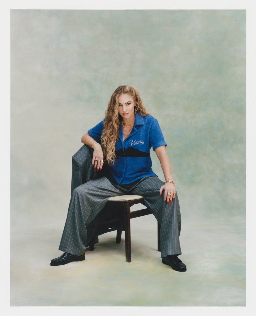 Female model with curly long blonde hair sitting facing the camera with one hand on her knee and the other resting against the backside of the chair in 'The Sopranos' Vesuvio Cabana ~ Crop Cut while wearing grey and white-striped baggy suit pants and balck loafers with a matching grey and white-striped blazer resting on the chair's backside, all of which was shot on a very light green background.