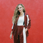 Female model with long blonde hair wearing 'The Sopranos' Pie-O-My Track Pants with a white shirt and a matching Pie-O-My Jacket, shot a textured red background.