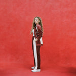 Female model with long blonde hair turned to the side while wearing 'The Sopranos' Pie-O-My Track Pants with matching Pie-O-My Jacket, a white shirt, and white sneakers, all shot on a red textured background.