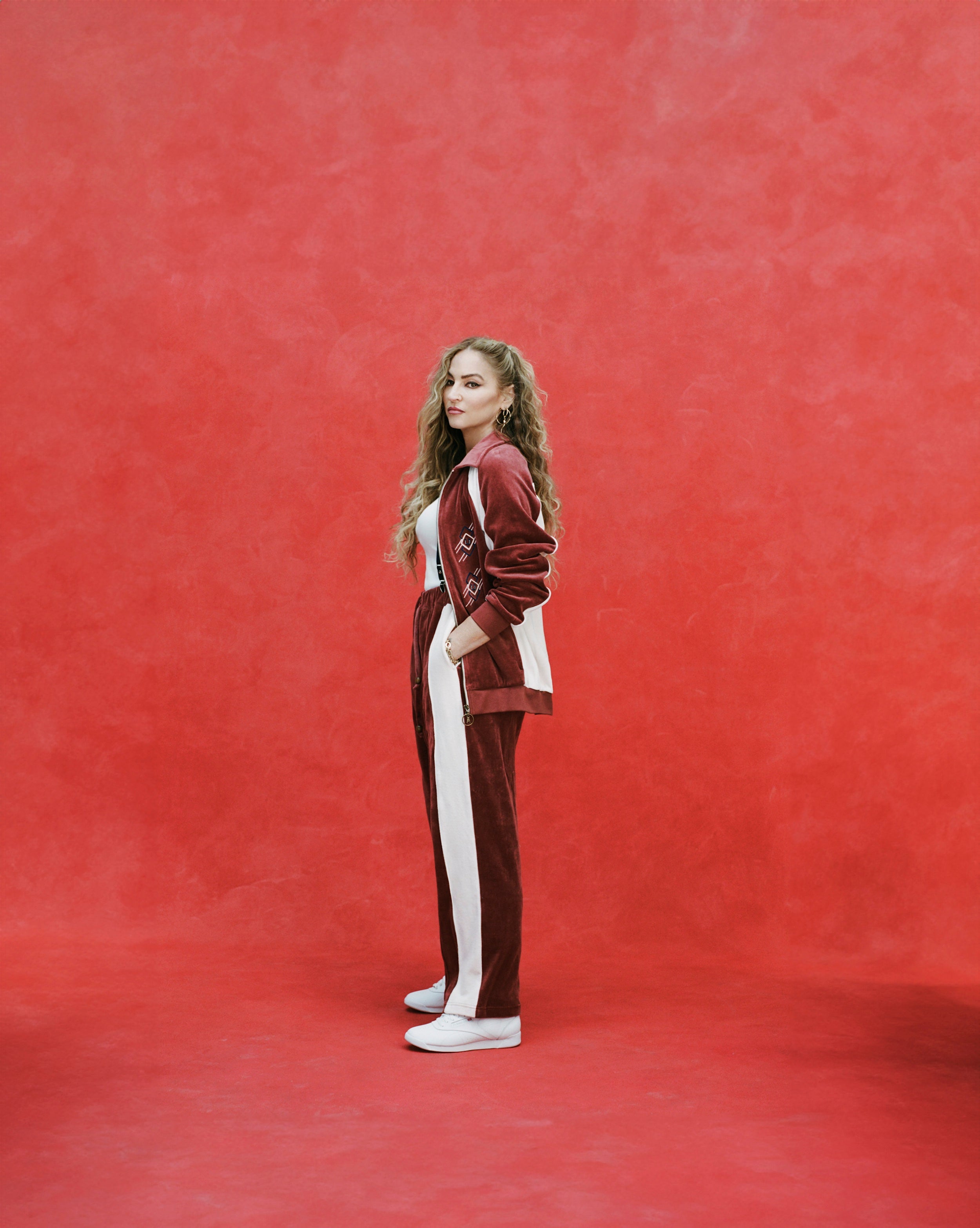 Female model with long blonde hair turned to the side while wearing 'The Sopranos' Pie-O-My Track Pants with matching Pie-O-My Jacket, a white shirt, and white sneakers, all shot on a red textured background.