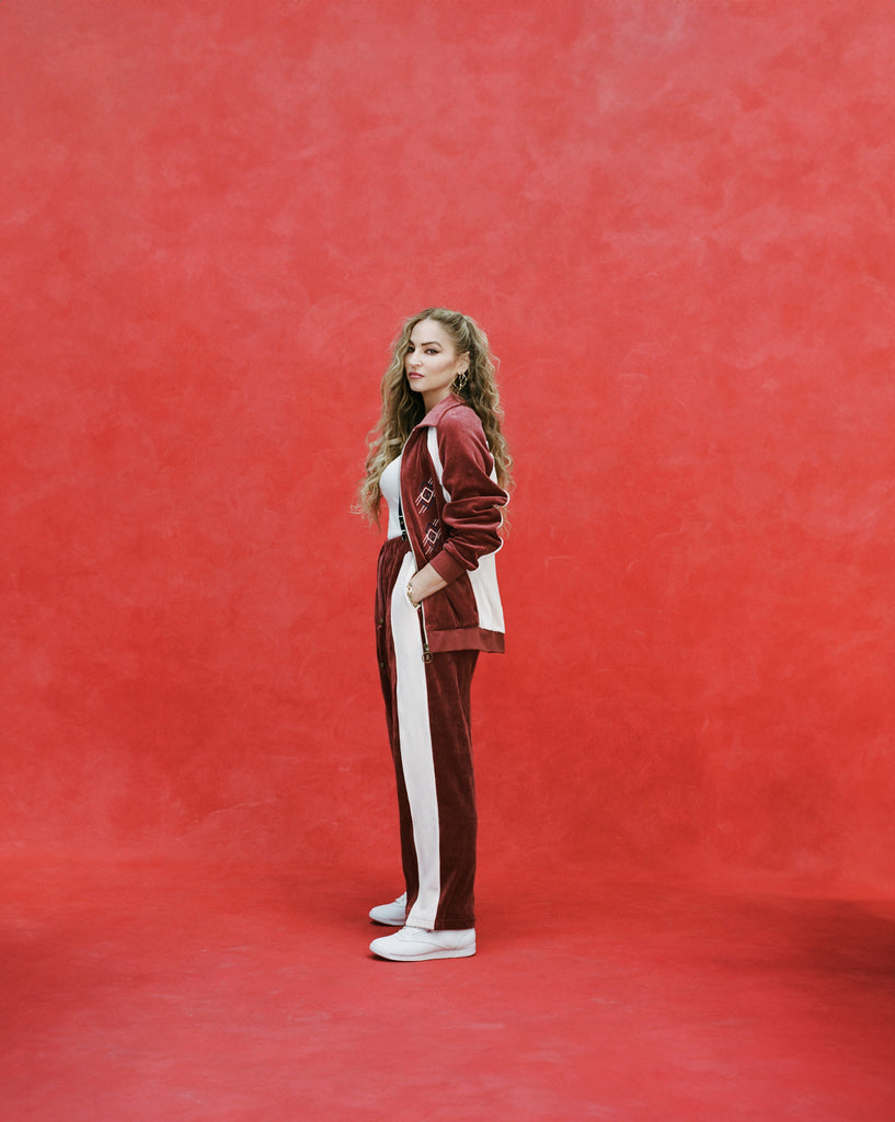 Female model with long blonde hair turned to the side while wearing 'The Sopranos' Pie-O-My Track Pants with matching Pie-O-My Jacket, a white shirt, and white sneakers, all shot on a red textured background.