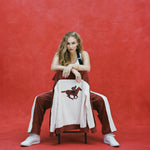 Female model with long blonde hair seated with the chair facing towards the camera and 'The Sopranos' Pie-O-My Jacket draped on it so that the chain stitched racehorse with jockey is prominently displayed, as the model is also wearing matching 'The Sopranos' Pie-O-My pants and white sneakers, shot on a red textured background.