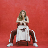 Female model with long blonde hair seated with the chair facing towards the camera and 'The Sopranos' Pie-O-My Jacket draped on it so that the chain stitched racehorse with jockey is prominently displayed, as the model is also wearing matching 'The Sopranos' Pie-O-My pants and white sneakers, shot on a red textured background.