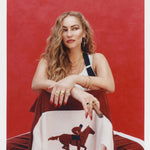 Female model with long blonde hair holding a cigar seated in a chair that faces the camera so that 'The Sopranos' Pie-O-My Track Jacket and its chain stitched racehorse hanging off of said chair is prominently displayed, she's also wearing a white tank top with suspenders, and matching 'The Sopranos' Pie-O-My Track Pants, all shot on a red textured background.