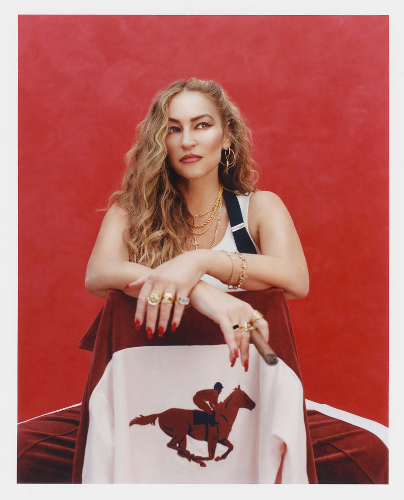 Female model with long blonde hair holding a cigar seated in a chair that faces the camera so that 'The Sopranos' Pie-O-My Track Jacket and its chain stitched racehorse hanging off of said chair is prominently displayed, she's also wearing a white tank top with suspenders, and matching 'The Sopranos' Pie-O-My Track Pants, all shot on a red textured background.