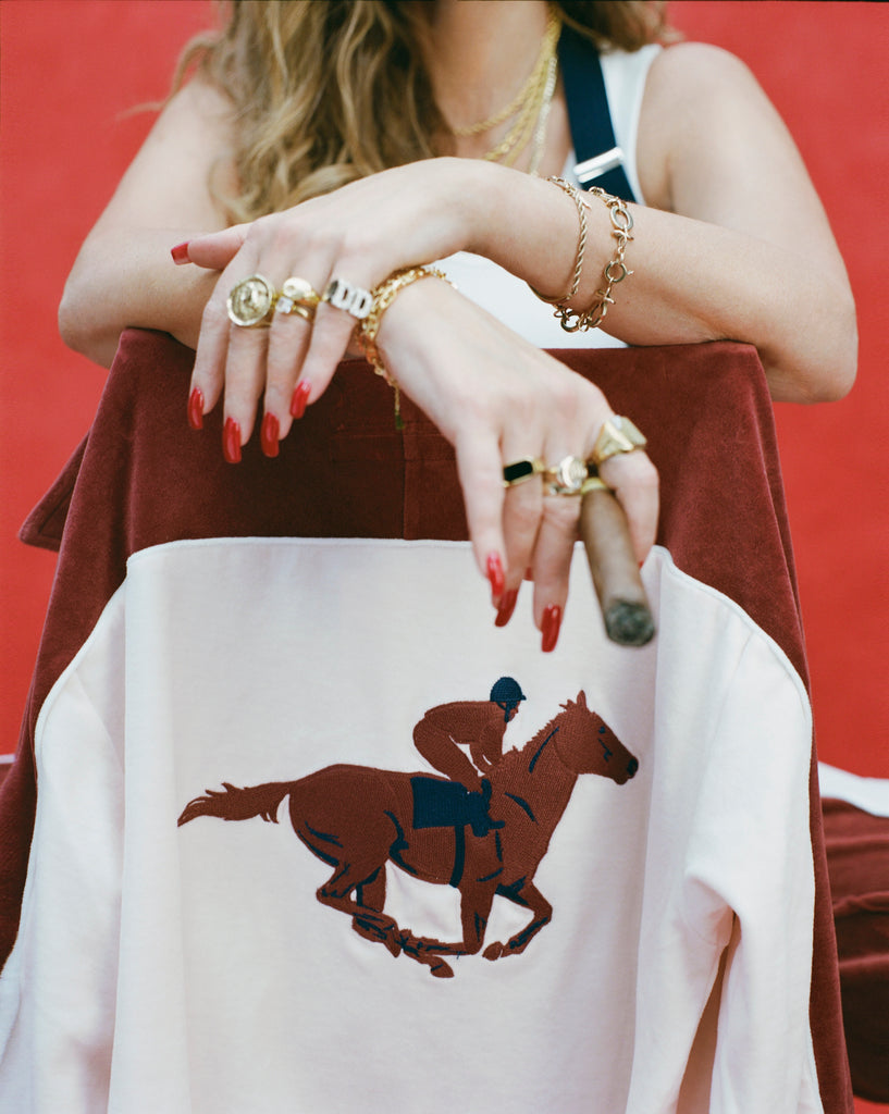 Close-up product image of 'The Sopranos' Pie-O-My Track Jacket draped over the back of a chair with the models hands hanging over it, holding a cigar and featuring gold jewelry and rings.