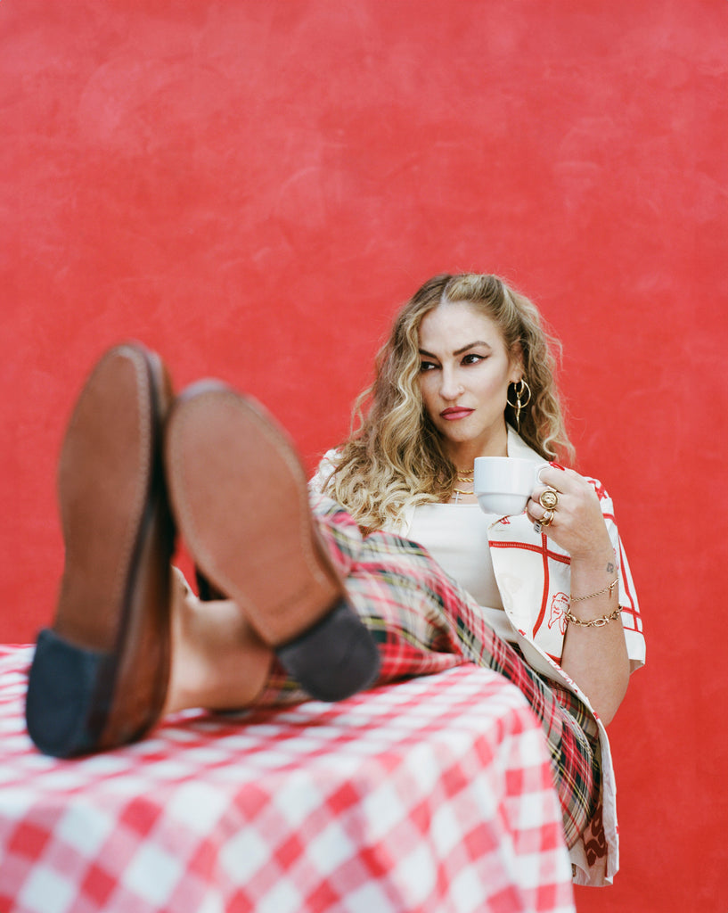 Female model with long blonde hair holding a coffee mug with her feat up on a table, wearing 'The Soprnaos' Satriale's (Gabagool) unbuttoned with a white shirt and plaid pants, on a red background.