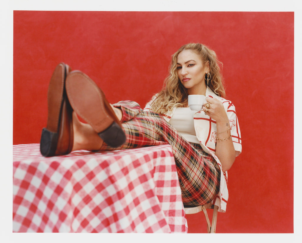 Female model with long blonde hair seated with a coffee mug in one hand, wearing red plaid pants, brown loafers, and 'The Sopranos' Satriale's (Gabagool) unbuttoned with a white shirt.