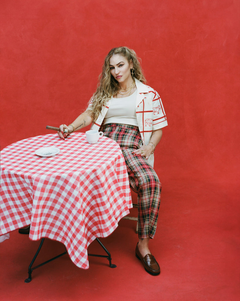 Female model with long blonde hair wearing an unbuttoned 'The Sopranos' Satriale's (Gabagool) seated with a cigar in one hand and the other hand on her leg at a table with a coffee mug, wearing plaid plants, white shirt, and brown loafers.