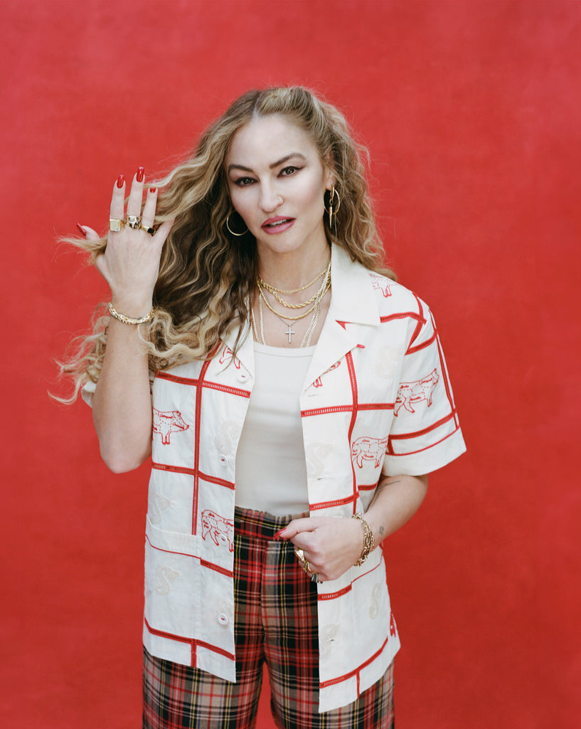 Female model with long blonde hair wearing 'The Sopranos' Satriale's (Gabagool) unbottoned with a white shirt, red plaid paints, on a red background, with one hand elevated.