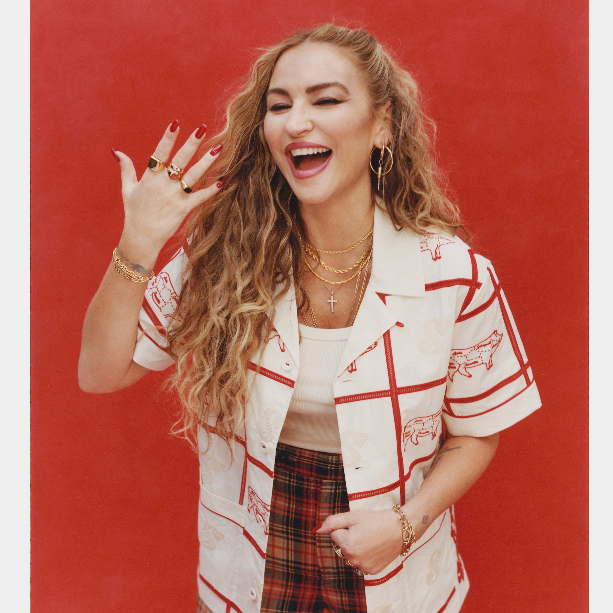 Female model with long blonde hair wearing an unbuttoned 'The Sopranos' Satriale's (Gabagool) that features a cream base, red stitching, paneling, and pig embroidery on alternating panels, labeled with various cuts in Sopranos parlance: "gabagool", "pruh-zhoot", etc. She is wearing plaid pants, and a white shirt, holding one hand up while laughing and wearing multiple gold rings, shot on a red background.
