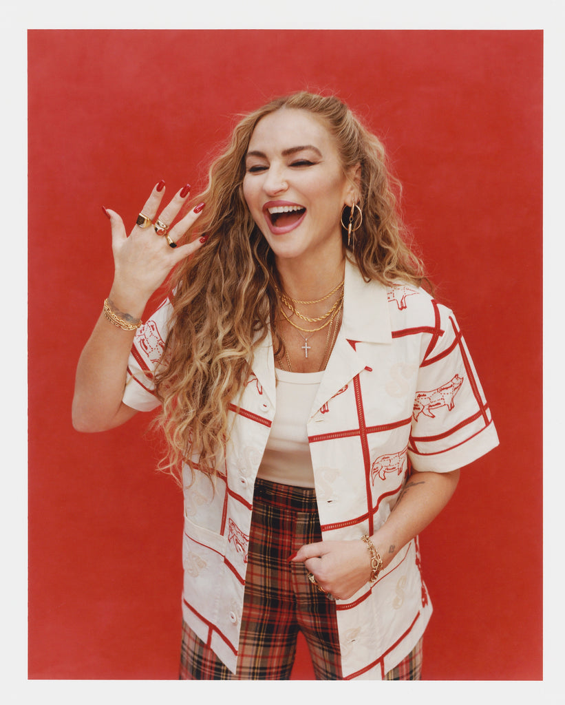 Female model with long blonde hair wearing an unbuttoned 'The Sopranos' Satriale's (Gabagool) that features a cream base, red stitching, paneling, and pig embroidery on alternating panels, labeled with various cuts in Sopranos parlance: "gabagool", "pruh-zhoot", etc. She is wearing plaid pants, and a white shirt, holding one hand up while laughing and wearing multiple gold rings, shot on a red background.