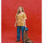 Female model with long blonde hair wearing 'The Sopranos' Satriale's (Pruh-Zhoot) while wearing baggy jeans, brown loafers, and holding a Houndstooth brown blazer and denim hat, all shot on a red background.