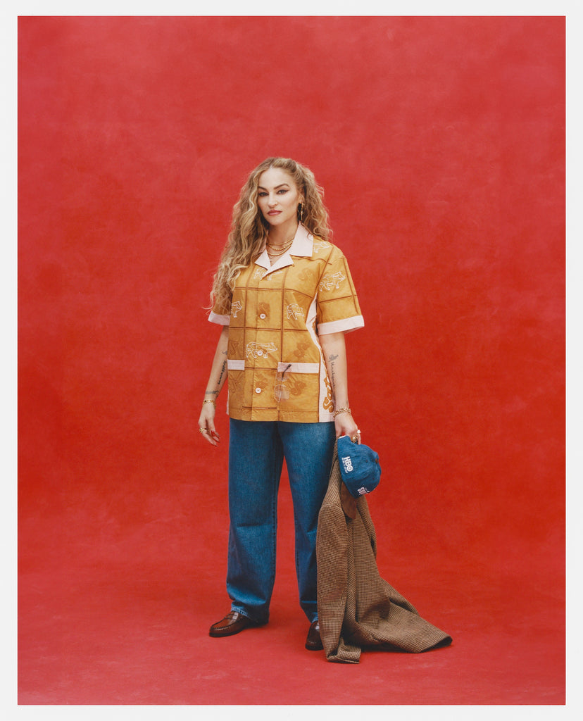 Female model with long blonde hair wearing 'The Sopranos' Satriale's (Pruh-Zhoot) while wearing baggy jeans, brown loafers, and holding a Houndstooth brown blazer and denim hat, all shot on a red background.