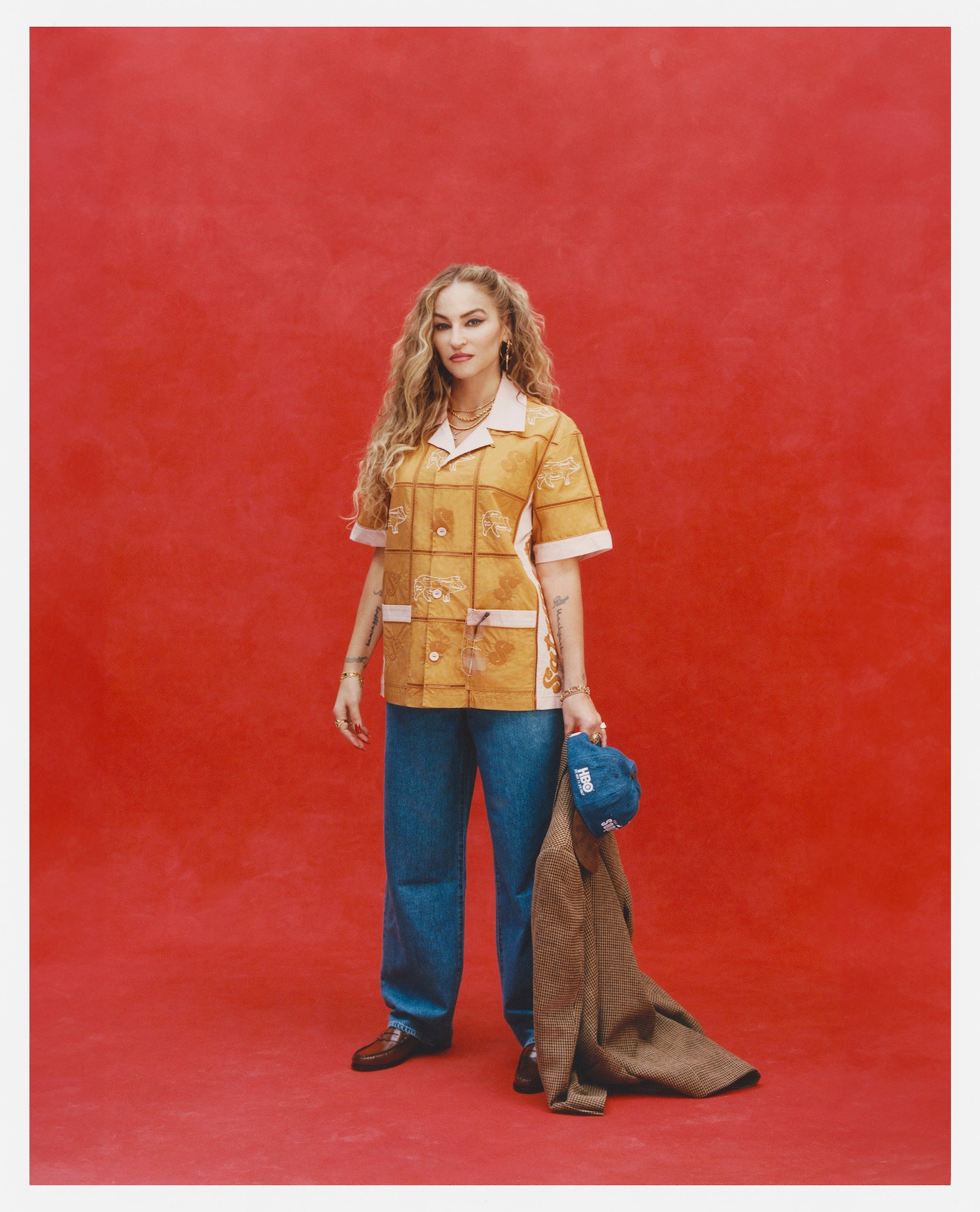 Female model with long blonde hair holding a Houndstooth blazer and 'The Sopranos' Fowl Play 6-Panel Souvenir Cap while wearing a Satriale's "Pruh-Zhoot" Cabana, long flowey jeans, and brown loafers, shot on a red textured background.