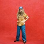 Female model with long blonde hair with arms at her waist wearing 'The Sopranos' Satriale's (Pruh-Zhoot) with flowy jeans, brown loafers, and a denim hat with a brown suede brim that says 'Sopranos,' all shot on a red textured background.