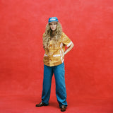 Female model with long blonde hair with arms at her waist wearing 'The Sopranos' Satriale's (Pruh-Zhoot) with flowy jeans, brown loafers, and a denim hat with a brown suede brim that says 'Sopranos,' all shot on a red textured background.