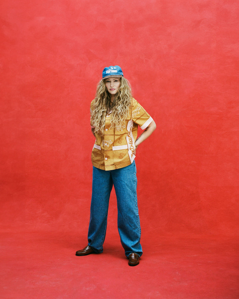 Female model with long blonde hair with arms at her waist wearing 'The Sopranos' Satriale's (Pruh-Zhoot) with flowy jeans, brown loafers, and a denim hat with a brown suede brim that says 'Sopranos,' all shot on a red textured background.