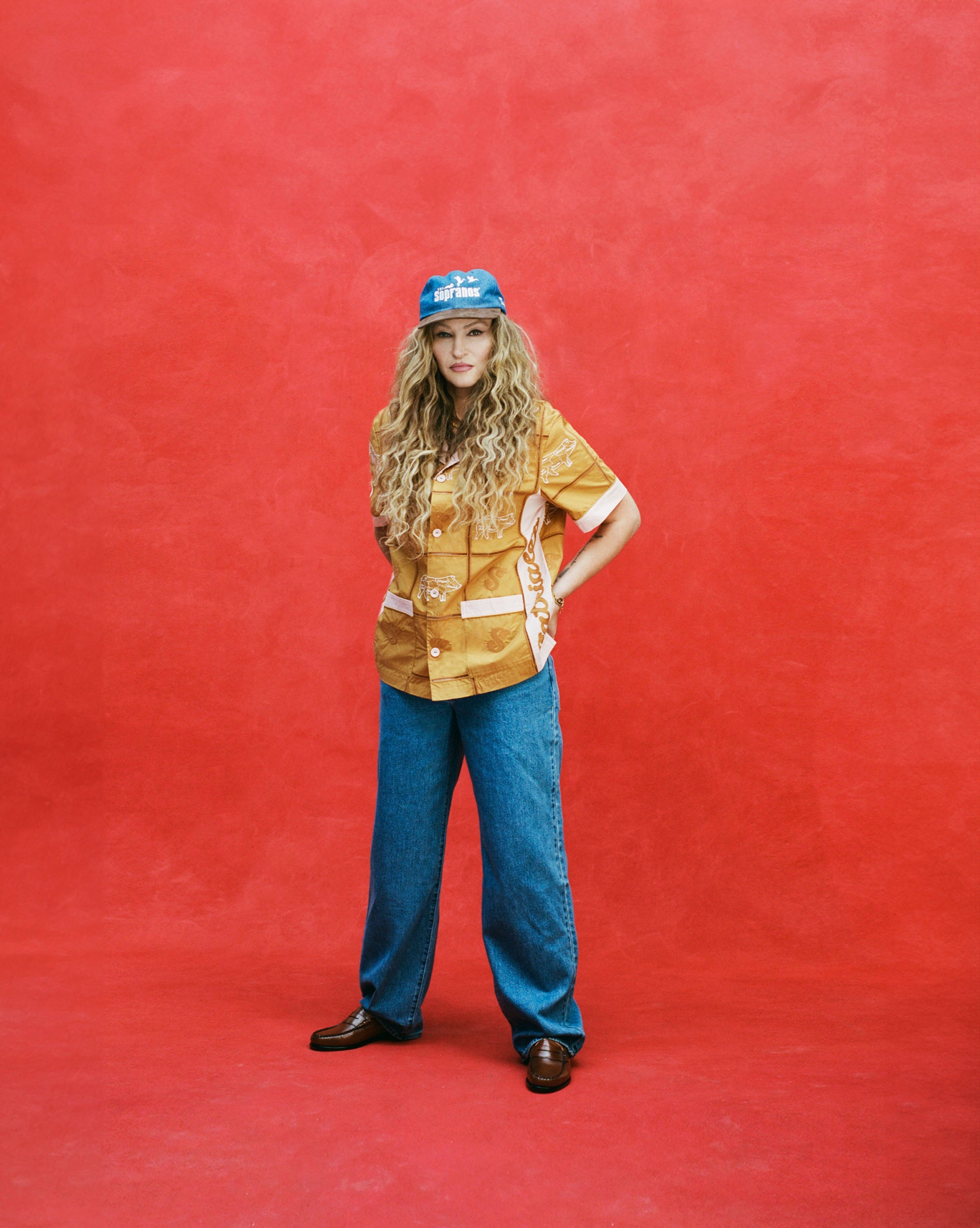 Female model with long blonde hair wearing 'The Sopranos' Fowl Play 6-Panel Souvenir Cap alongside a Satriale's 'Pruh-Zhoot' Cabana with long flowy jeans and brown loafers, shot on a textured red background.