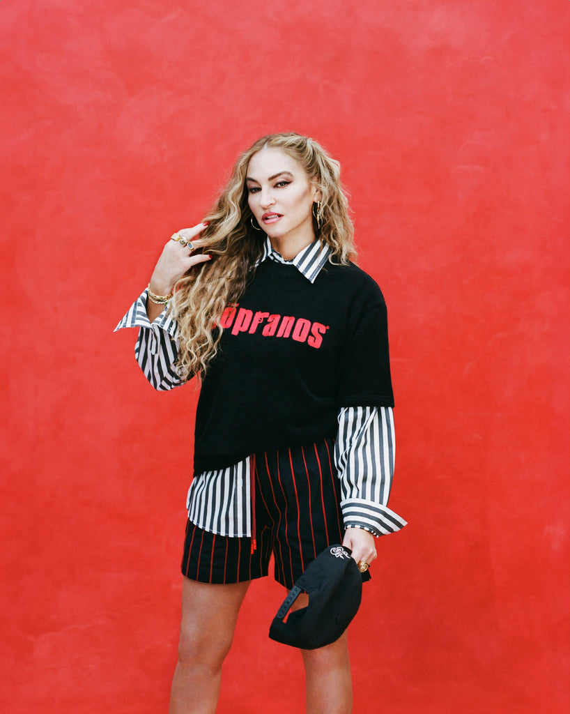 Female model with long blonde hair wearing 'The Sopranos' Lineup Swim Trunks with a Sopranos graphic terry tee, black and white striped collared shirt beneath that, while holding a black snap-back hat with her other hand running through her hair, shot on a red textured background.