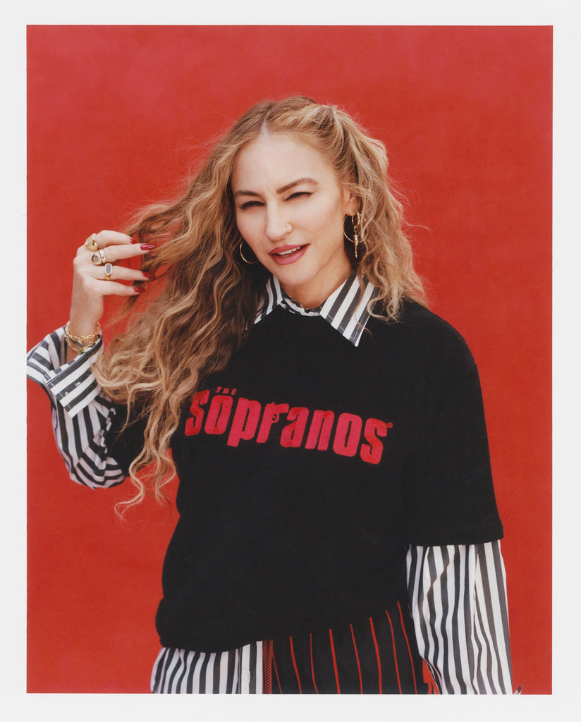 Female model with long, wavy blonde hair is smiling and slightly squinting at the camera; she is wearing the "The Sopranos" Souvenir Terry Tee over a black-and-white striped shirt, with a red background behind her.