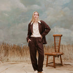 Female model with long blonde hair wearing the 'Easy-Going Jacket' brown linen unbuttoned with a white button down shirt with large collars, matching brown linen pants, and light brown loafers, shot on a background with light blue texturing, waist high beige grass, and a bamboo chiar.