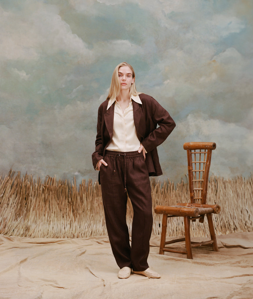 Female model with long blonde hair wearing the 'Easy-Going Jacket' brown linen unbuttoned with a white button down shirt with large collars, matching brown linen pants, and light brown loafers, shot on a background with light blue texturing, waist high beige grass, and a bamboo chiar.