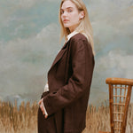Female model with long blonde hair turned away from the camera wearing the 'Easy-Going Jacket' in brown linen with matching pants in brown linen, shot on a light-blue textured background, waist-high beige grass, and a bamboo chair.