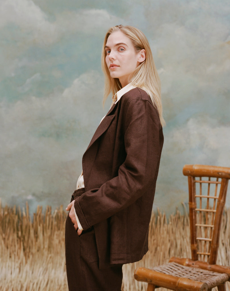 Female model with long blonde hair turned away from the camera wearing the 'Easy-Going Jacket' in brown linen with matching pants in brown linen, shot on a light-blue textured background, waist-high beige grass, and a bamboo chair.