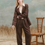 Female model with long blonde hair wearing the 'Easy-Going' Jacket in brown linen with a beige shirt with a large collar and matching brown linen jacket, and beige loafers, shot on a light blue textured background with waist high grass and a bamboo chair.