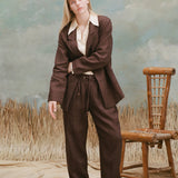 Female model with long-blonde hair wearing the 'Easy-Going' pants in brown linen alongside a brown blazer, cream colored shirt with oversized collars, unbuttoned half-way, and beige loafers, shot on a light-blue textured background with a bamboo chair and waist-high beige grass.