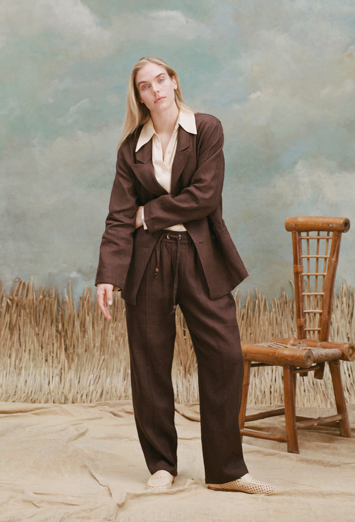 Female model with long-blonde hair wearing the 'Easy-Going' pants in brown linen alongside a brown blazer, cream colored shirt with oversized collars, unbuttoned half-way, and beige loafers, shot on a light-blue textured background with a bamboo chair and waist-high beige grass.