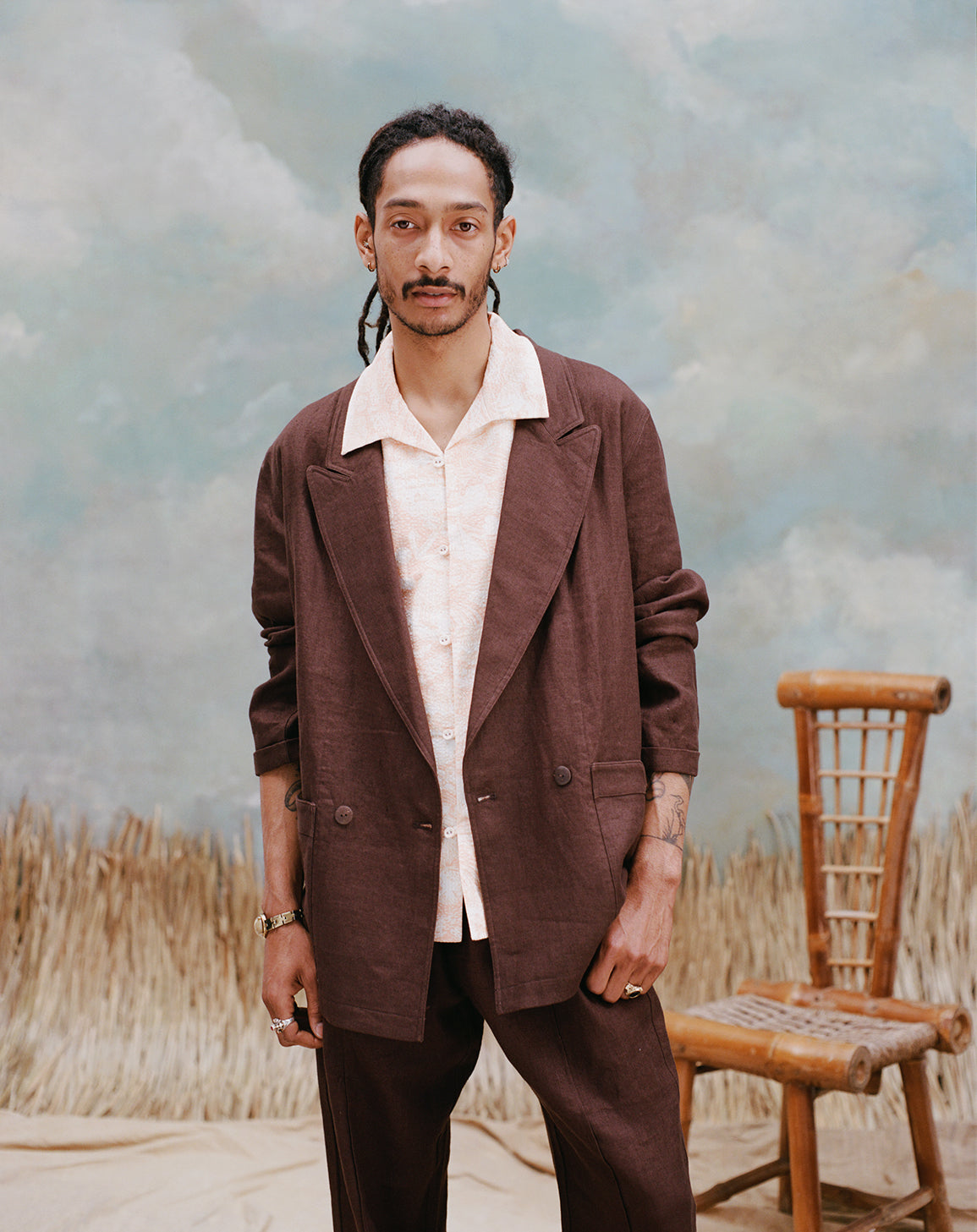 Male model with dreadlocks wearing the Voyager Shirt in coral seersucker beneath an Easy-Going jacket in brown linen with brown linen pants, shot on a light blue textured background with beige grass and bamboo chair.