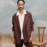 Male model with dreadlocks wearing the Voyager Shirt in coral seersucker beneath an Easy-Going jacket in brown linen with brown linen pants, shot on a light blue textured background with beige grass and bamboo chair.